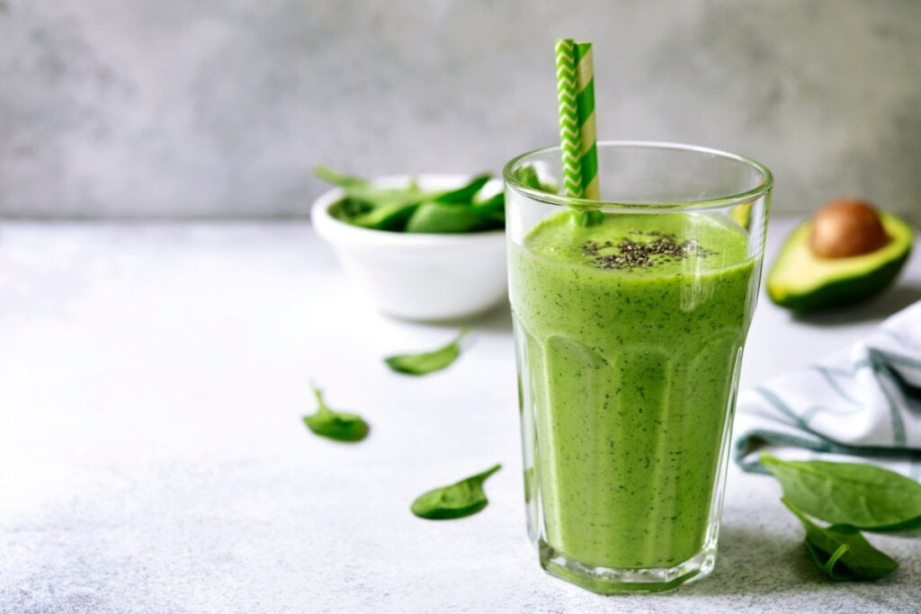 8 vitaminas caseiras para fazer no liquidificador 6 copo com vitamina de abacate e espinafre em cima de uma bancada e com espinafre em uma cumbuca e metade de um abacate atrás