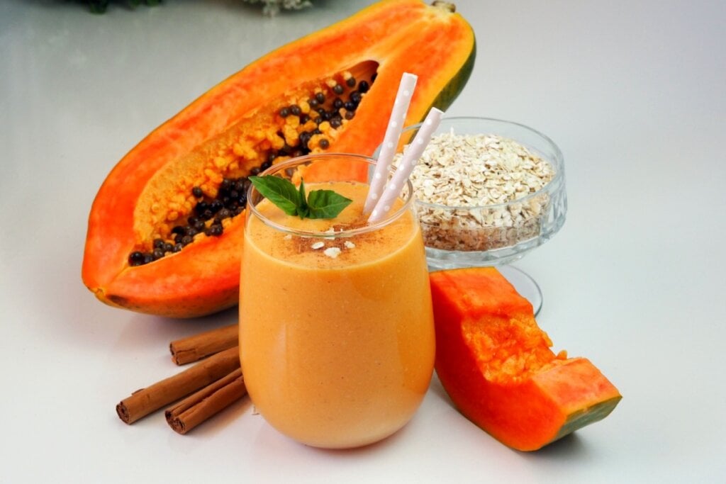 7 vitaminas termogênicas com canela para emagrecer com saúde 6 Vitamina de mamão com aveia e canela servido em copo de vidro em cima de mesa de mármore branco, ao lado está metade de um mamão, uma tigela com aveia e canela em pau