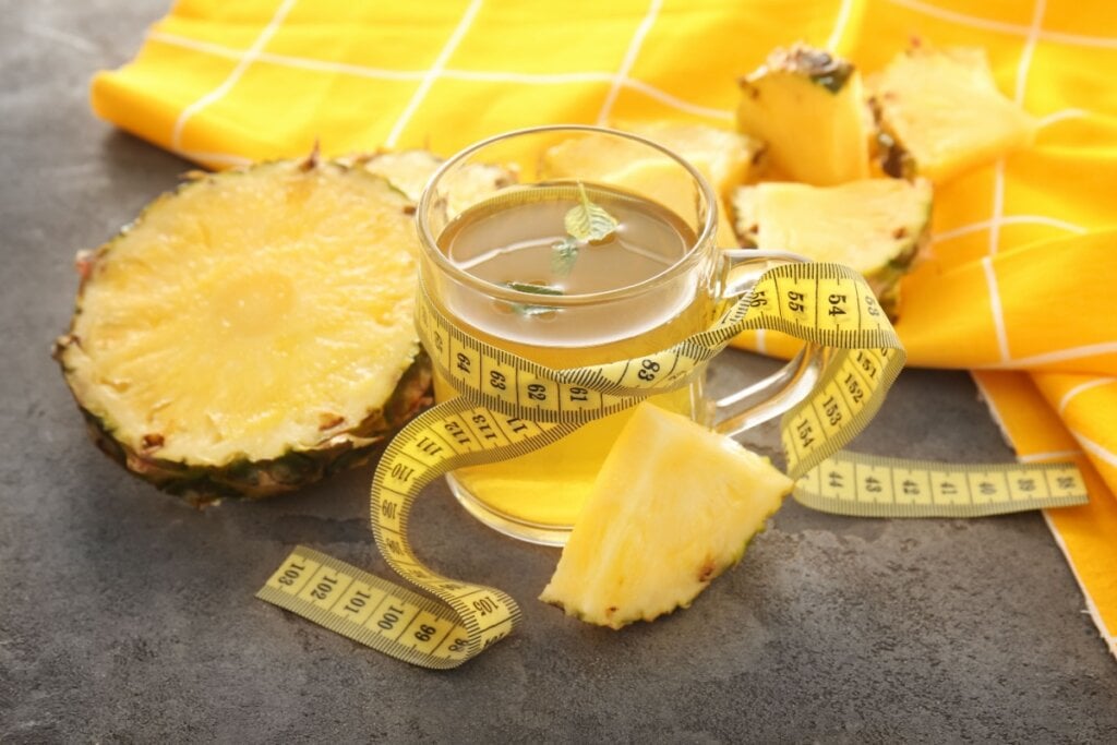 7 chás para ajudar a eliminar gordura abdominal 6 Xícara de chá com abacaxi, cercada por pedaços de abacaxi fresco e uma fita métrica