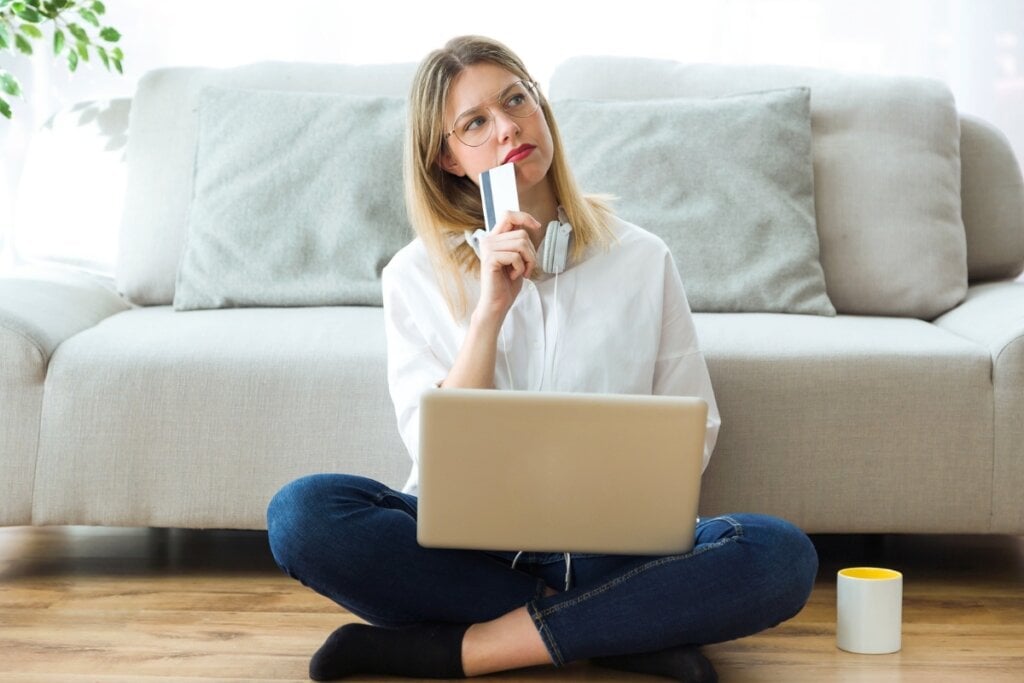 30 atitudes simples para preservar o meio ambiente 6 Mulher sentada com um notebook no colo, olhando para o lado e segurando um cartão próximo ao queixo como se estivesse pensando