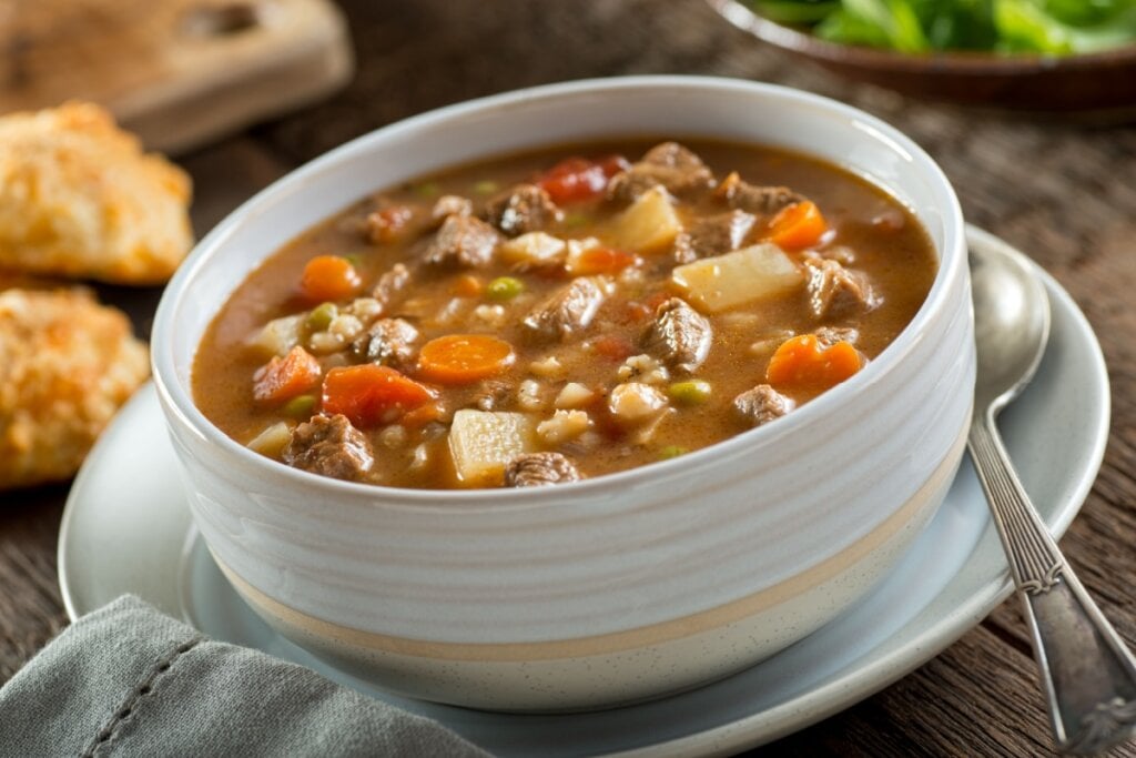 8 sopas ricas em proteínas para manter a dieta em dias frios 6 Sopa de carne com cevada e legumes em tigela de cores claras em cima de prato de mesma tonalidade
