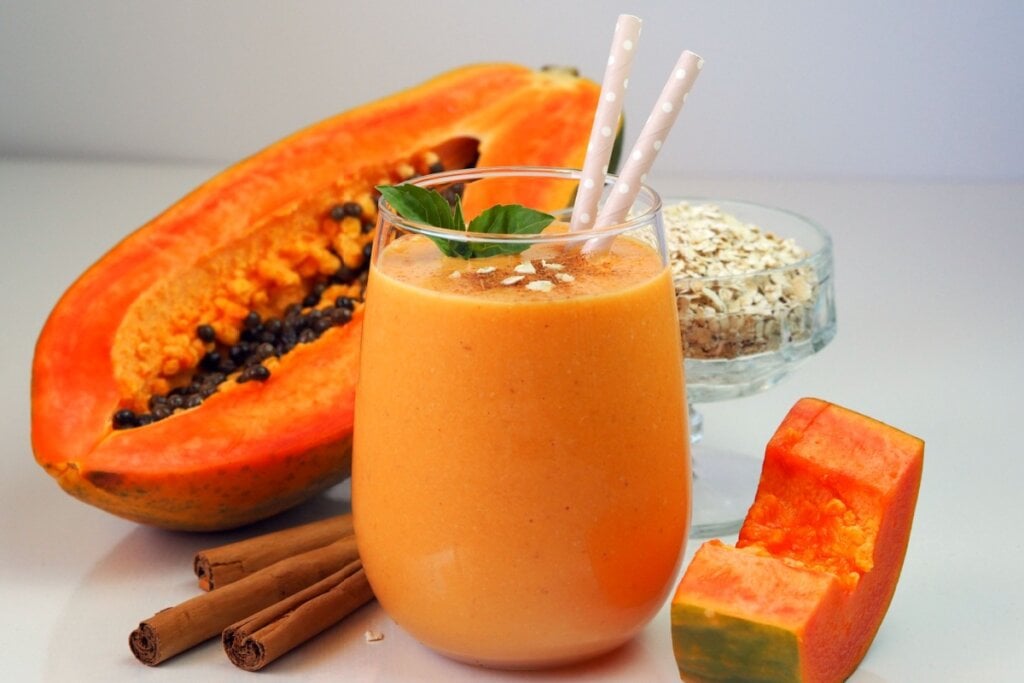 9 vitaminas com frutas para ajudar a emagrecer 6 Vitamina de mamão com aveia e canela servido em copo de vidro, decorado com hortelã e canudos, ao lado de mamão, pau de canela e aveia em flocos