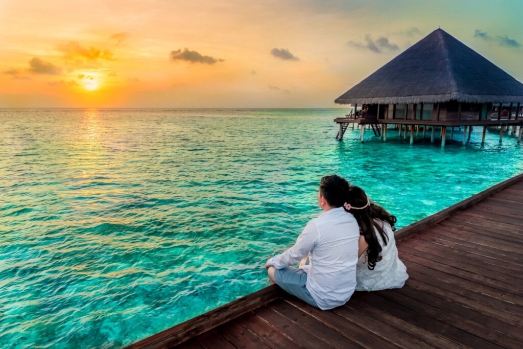 5 destinos românticos para visitar no Dia dos Namorados 24 Casal sentado em um deck de madeira assistindo ao pôr do sol diante do mar azul-turquesa nas Maldivas ao lado de bangalô