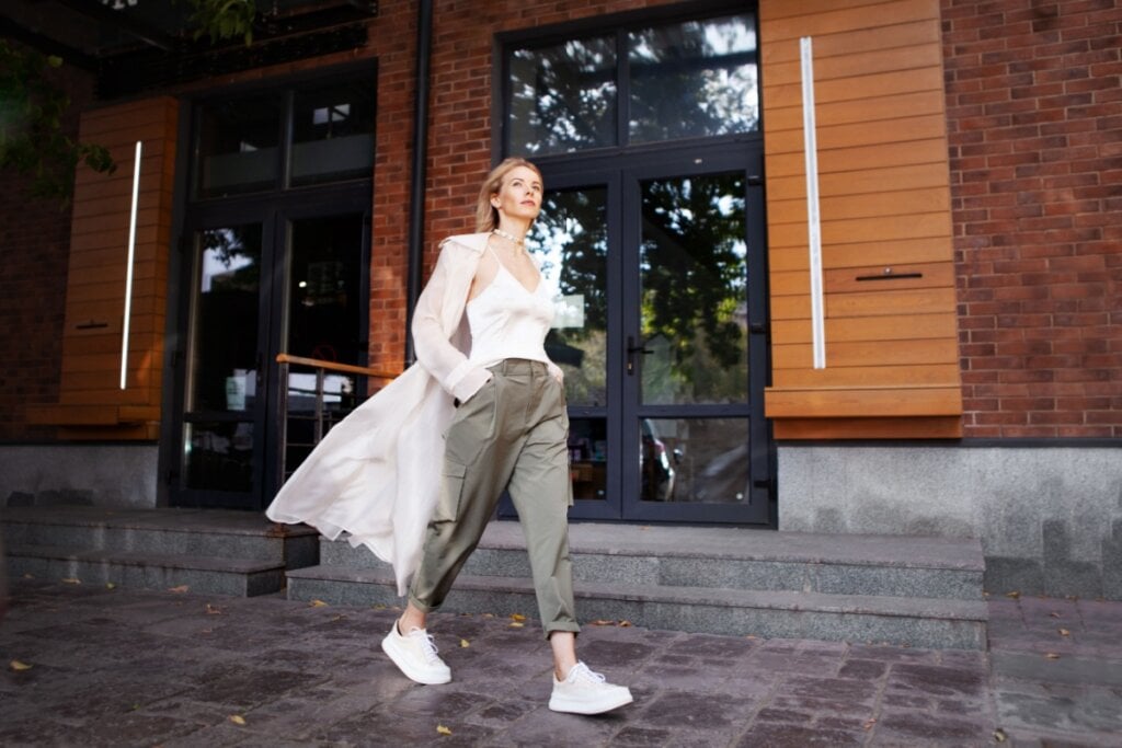 5 tendências de moda casual para o inverno 23 Mulher caminhando em frente a prédio de tijolos com calça cargo verde-musgo, regata branca e casaco esvoaçante
