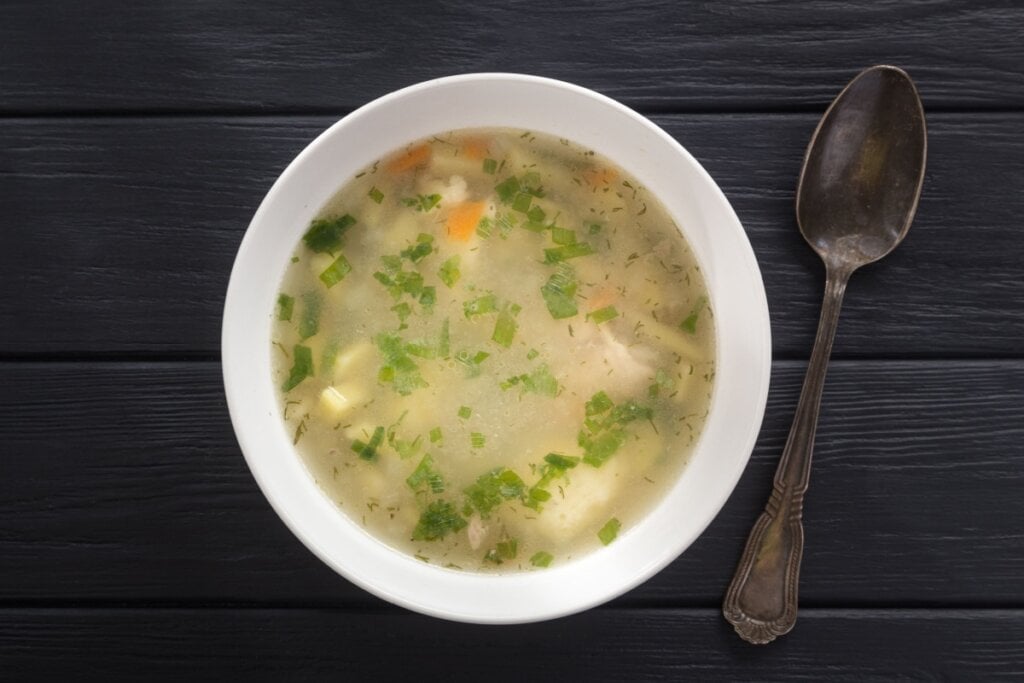 7 cremes e caldos para a festa junina 6 Caldo de frango servido em tigela de cerâmica branca, com colher ao lado em cima de mesa de madeira