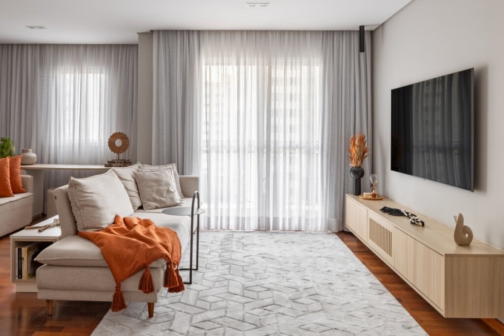 4 dicas práticas para aquecer a casa no inverno 16 Sala de estar com sofá claro decorado com almofadas e manta laranja, cortinas translúcidas e tapete geométrico sobre piso de madeira